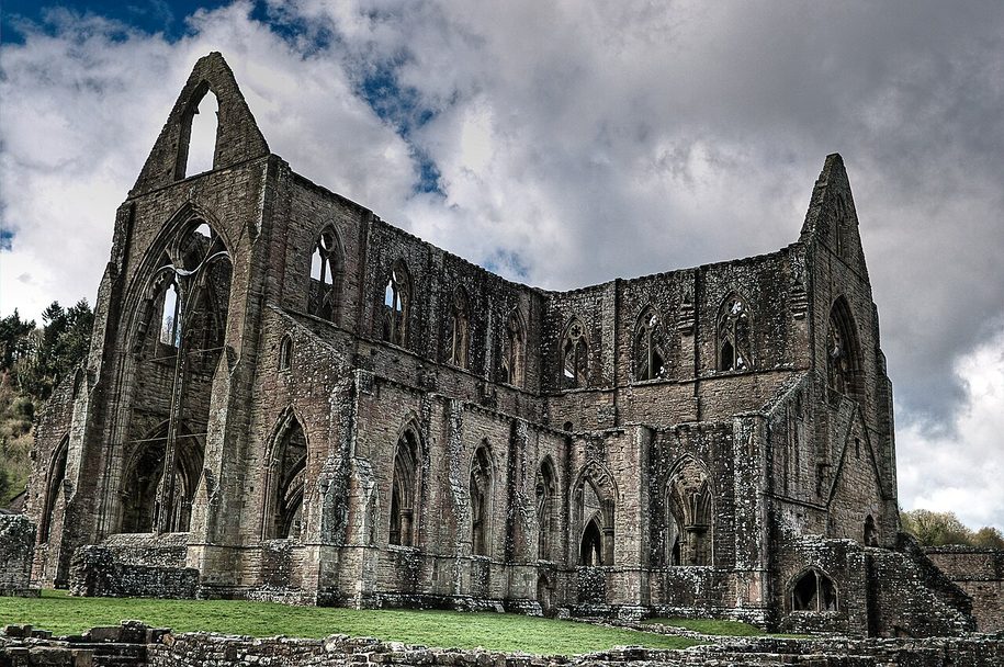 The full length of Tintern Abbey church under a cloudy sky, with the wooded slopes of the Wye Valley behind
