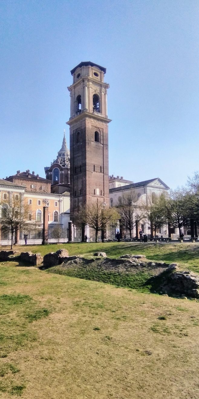 The tall brick bell tower of Turin Cathedral seen across the grass and ruins of the Roman theatre
