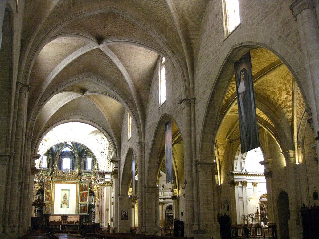 The Gothic nave of Valencia Cathedral with ribbed vaults and pale stone arches, leading to the brightly lit high altar