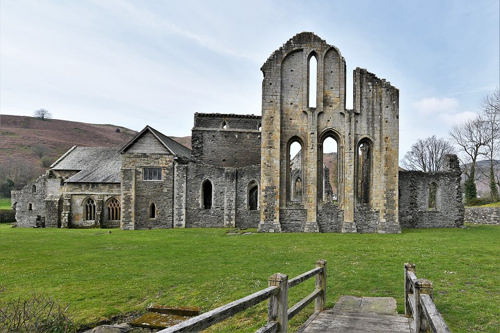 Valle Crucis Abbey near Llangollen: the tall ruined east end of the church beside the roofed east range, on a green lawn below wooded hills