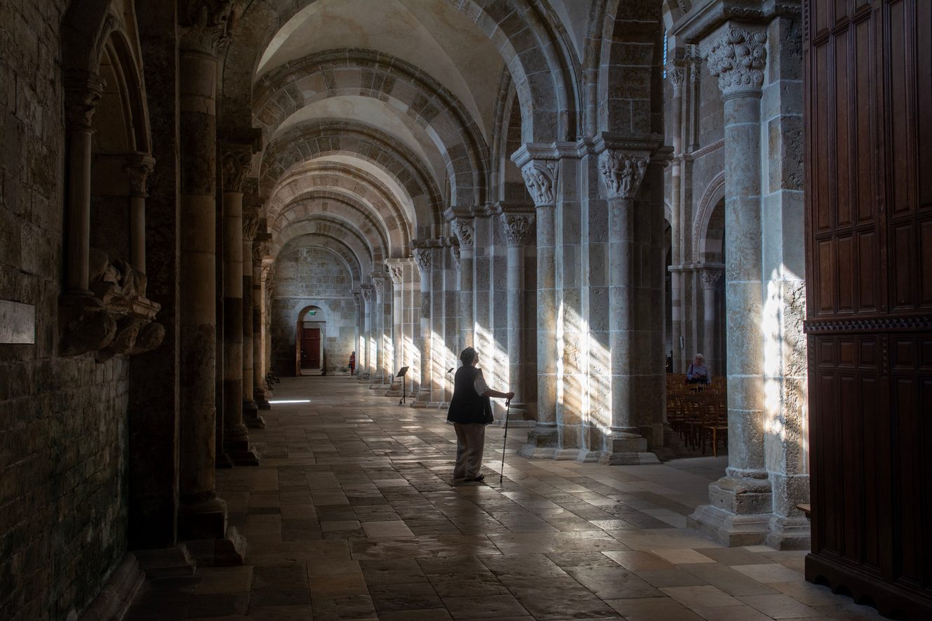 Sunlight falling through the arches of a side aisle at Vézelay onto the stone floor, with a visitor walking in the light