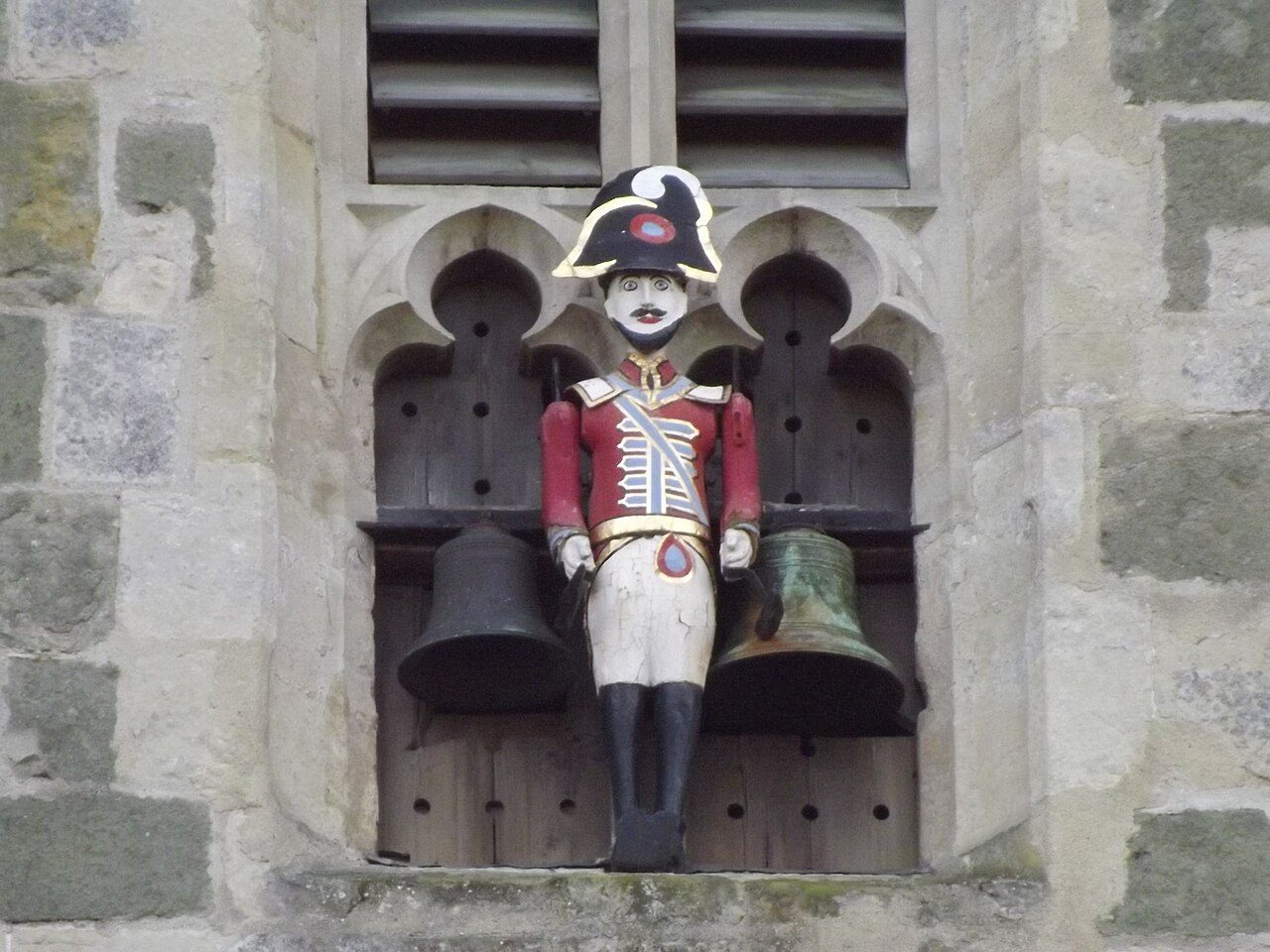 A painted wooden figure of a soldier in a red coat standing between two bells on the tower
